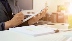 business person reviewing data on a clipboard with tablet and phone on table next to stack of papers business person reviewing data on a clipboard with tablet and phone on table next to stack of papers