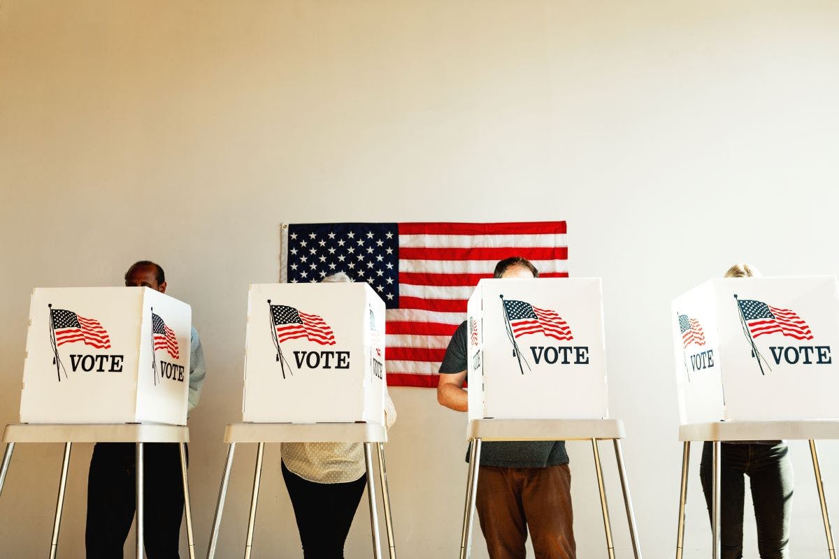 U.S. election day; people at voting booth at U.S. election station with American flag in background.