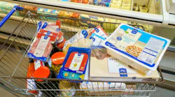 A shopping cart full of food items including pizza, cheese, and meat. The cart is in a store and the items are in the freezer section.