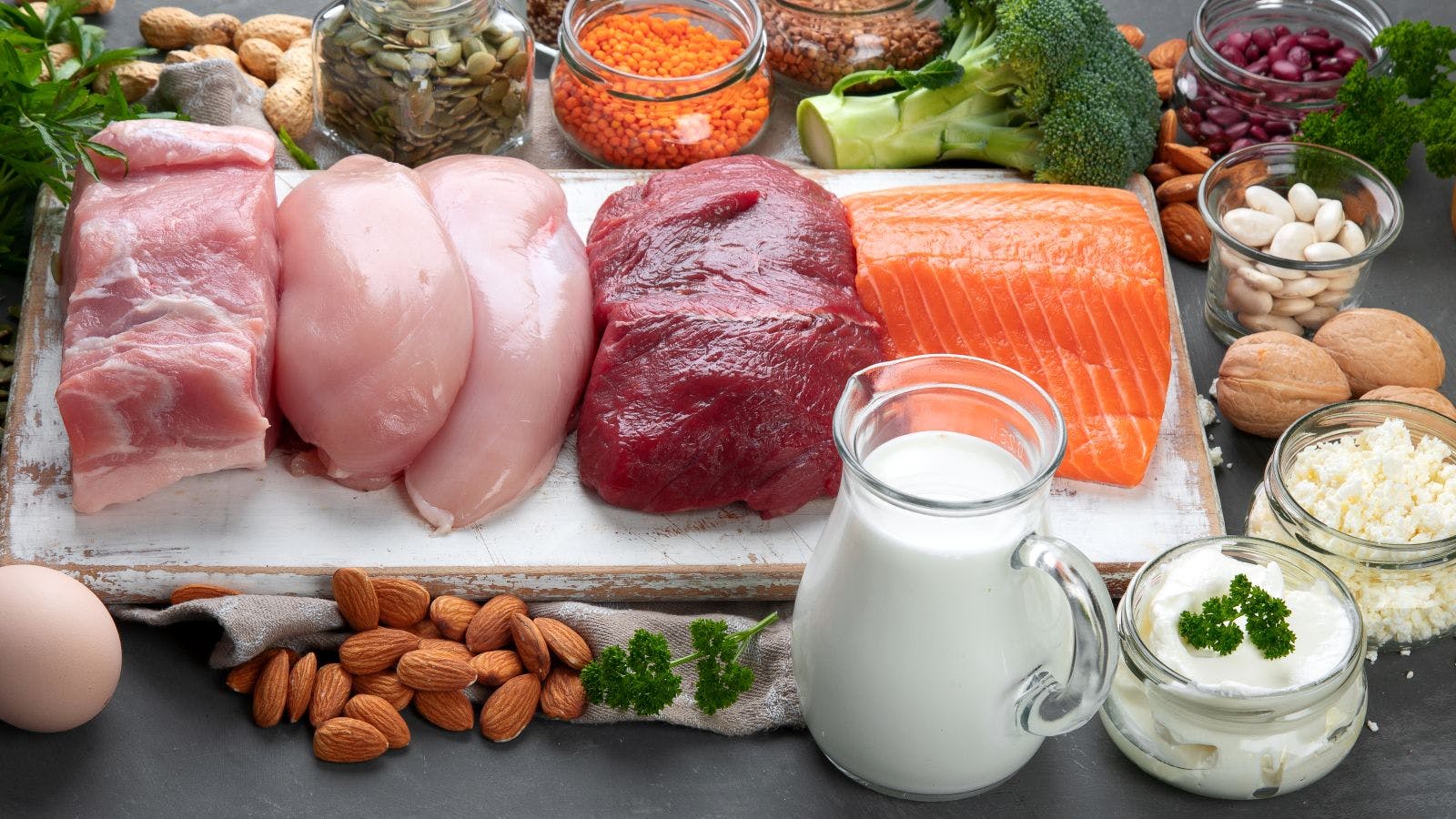 An array of food laid out across a table: raw pork, raw chicken, raw steak, raw salmon, a pitcher of milk, a handful of almonds, broccoli, walnuts, cream cheese, crumbly feta cheese, and more.