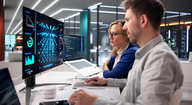 Two specialists oversee real-time server inference operations in data center environment using laptops.
