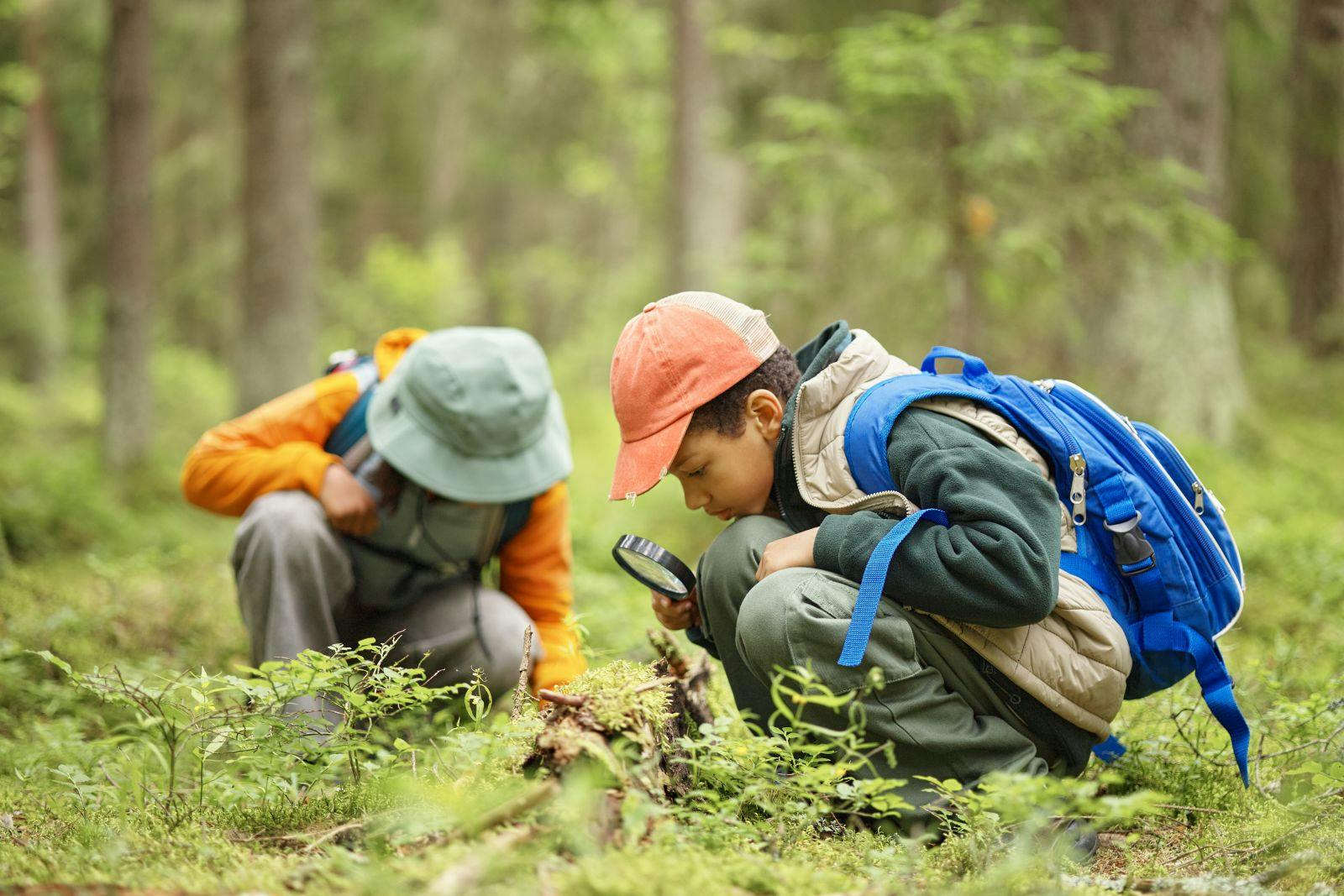 Two children exploring forest floor, one boy with blue backpack using magnifying glass to examine plants while another child in background observing nature closely.