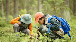 Two children exploring forest floor, one boy with blue backpack using magnifying glass to examine plants while another child in background observing nature closely.