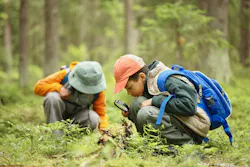 Two children exploring forest floor, one boy with blue backpack using magnifying glass to examine plants while another child in background observing nature closely.