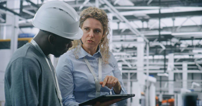 Two Specialists Solve Technical Issue in Vast Industrial Plant. Woman Points Area Concern on Factory Floor, Using Tablet For Data Reference. Problem Solving and Operational Excellence in Industry 4.0.